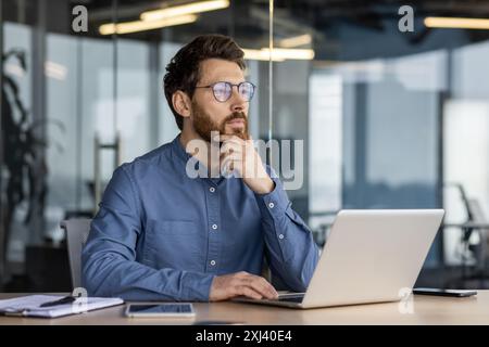 A serious and pensive young man is sitting in the office at a desk with a laptop, holding his head with his hand and looking ahead in concentration. Stock Photo
