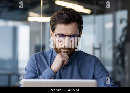 Close-up photo of a serious young businessman, programmer and developer sitting in the office at the desk, thoughtfully and intently looking at the laptop screen. Stock Photo
