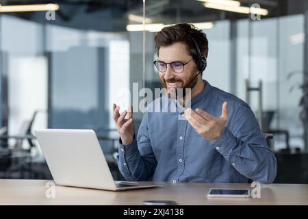 Smiling young man talking on a video call on a laptop, sitting in the office in a headset and gesturing with his hands to explain. Stock Photo