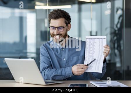A smiling young man works in the office, sits at a desk in front of a laptop screen and shows data on documents with a pen, conducts an online business meeting. Stock Photo