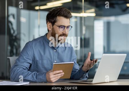 A smiling young man is sitting in the office, holding a notebook and talking on a video call, gesturing with his hands. Stock Photo