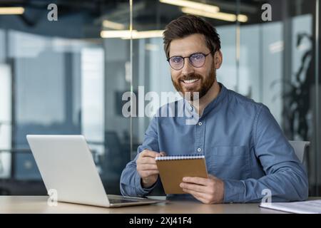 Portrait of a young smiling man in glasses working and studying in the office, sitting at a table with a notebook, holding a notebook with a pen, looking at the camera. Stock Photo