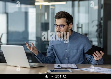 Frustrated businessman sitting at desk with laptop and tablet, displaying confusion and stress. Concept of workplace challenges, multitasking, and problem-solving in modern office environment Stock Photo