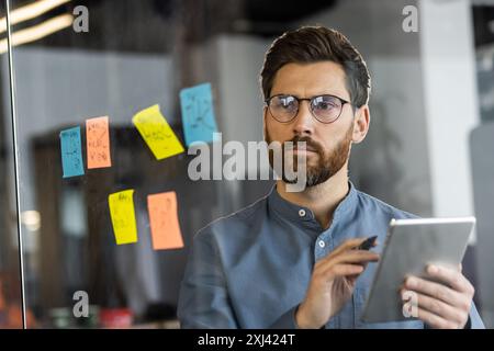 Close-up photo of a young man with glasses and a beard standing in the office near a transparent board, with colored sticky notes and writing data on a tablet. Stock Photo