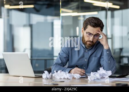 Upset and discouraged young man sitting in the office at the desk, holding his head with his hand, looking thoughtfully and disappointedly at the crumpled paper on the desk. Stock Photo