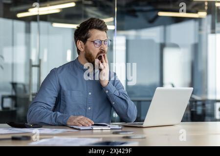 Tired young man, office worker sits in the office overtime, works on a laptop, yawns, covers his mouth with his hand. Stock Photo