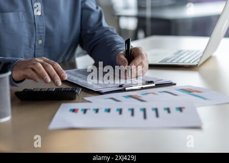 Business professional analyzing financial charts and graphs at desk. Hands holding pen and documents, calculator and laptop in background. Concept of data analysis, finance, corporate performance Stock Photo