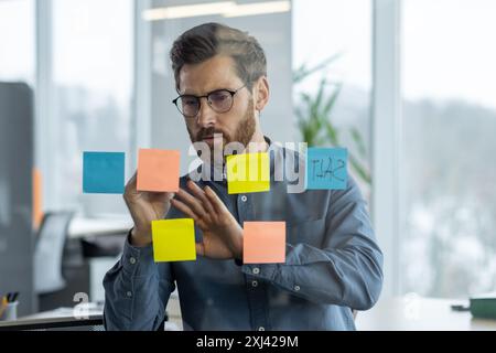 Close-up photo of a serious and focused young man working in an office, standing near a transparent information board and making notes on colored sticky notes. Stock Photo