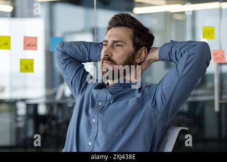Confident business professional relaxing in modern office with colorful sticky notes on glass wall. Person in blue shirt looking thoughtful, representing relaxation, contemplation Stock Photo