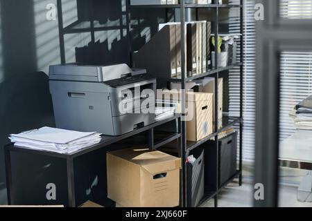 Plastic copier machine standing on desk with stack of paper documents by stand with shelves with cardboard boxes and folders with bills Stock Photo