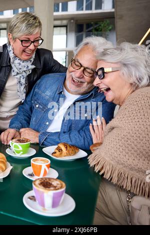 Three mature grey hair friends laughing together enjoying snacks sitting at cafeteria table.  Stock Photo