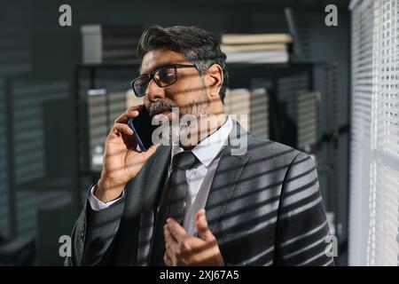 Bearded mature lawyer in eyeglasses and grey suit standing in front of camera in office and speaking with client on mobile phone Stock Photo