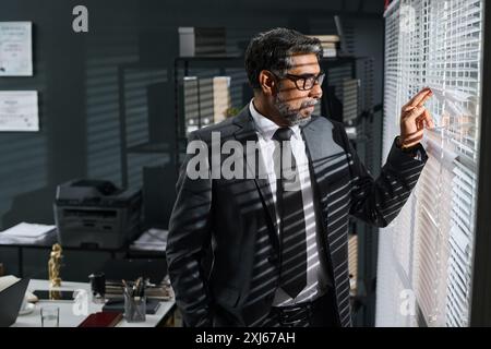 Side view of mature male lawyer or small business owner in classy suit looking through venetian blinds in office on sunny day Stock Photo