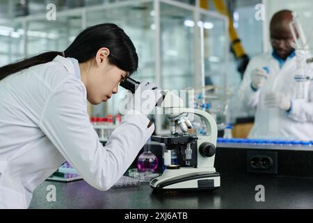 Side view of young Chinese female biochemist in whitecoat looking in microscope while studying new virus in chemical laboratory Stock Photo