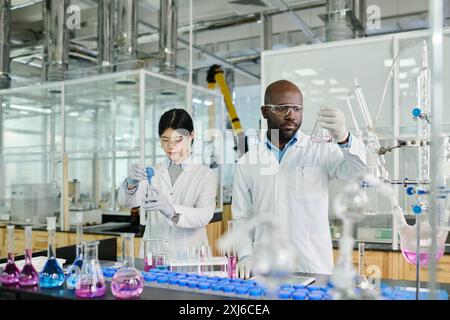 Young serious male scientist in lab coat looking at liquid in beaker while standing next to female researcher or chemist during experiment Stock Photo