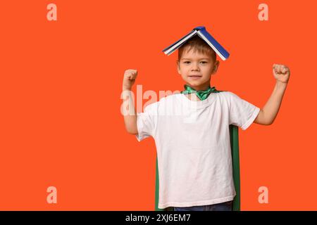 Cute little boy dressed as superhero with book on orange background. Library Lovers Day Stock Photo