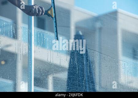 Close-up view of a hand using a squeegee to clean a glass balustrade, capturing cascading water drops and reflections of a contemporary building. Idea Stock Photo