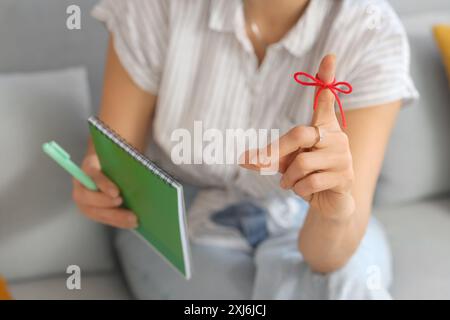 Woman with red string on her finger writing something in notebook at home, closeup. Reminder concept Stock Photo