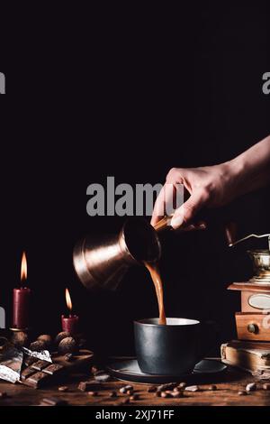cropped shot of woman pouring coffee from turk into cup at table with chocolate, truffles, candles and coffee grains on black background Stock Photo