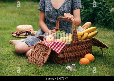 cropped image of woman sitting on green grass at picnic and touching wicker basket Stock Photo