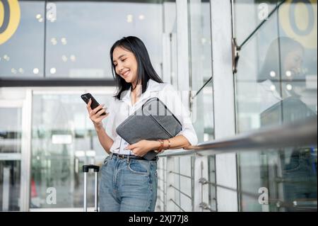 An attractive Asian female passenger is using her smartphone while standing at the exit door of the airport terminal with her belongings, waiting for Stock Photo