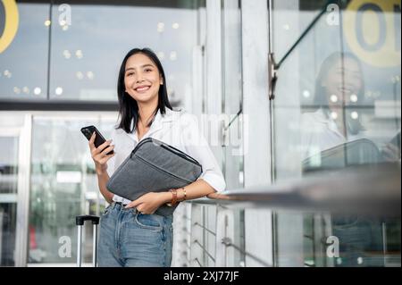 An attractive Asian female passenger with a smartphone in her hand is standing at the exit door of the airport terminal with her belongings, smiling a Stock Photo