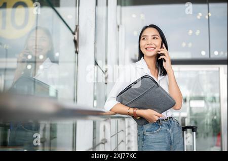 An attractive Asian female passenger is calling someone, talking on the phone while standing at the exit door of the airport terminal with her belongi Stock Photo