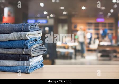 Blue denim jeans stack on wood table top with abstract blur clothing store in modern shopping mall defocused bokeh light background Stock Photo