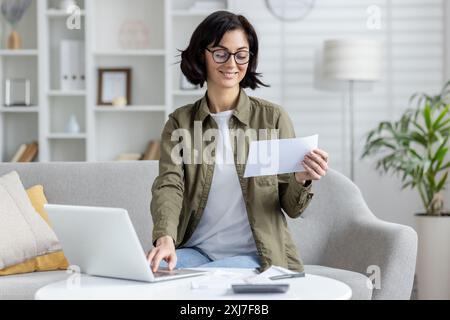 Smiling young woman sitting on sofa at home and working on notebook with documents, using laptop and holding papers and bills in hands. Stock Photo