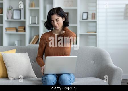 Photo of a young woman sitting at home on the sofa, holding a laptop on her lap, working and studying remotely, closing her eyes, massaging her neck with her hand, feeling severe pain and tension. Stock Photo