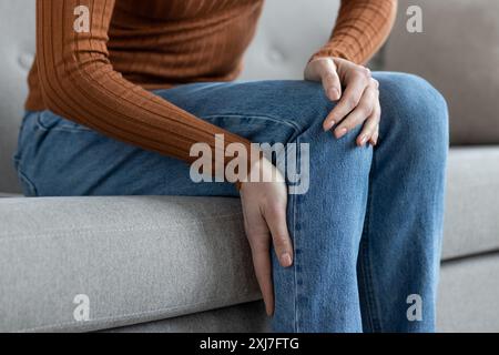Close-up of a woman in jeans and brown top sitting on a couch, holding her knee in discomfort. Stock Photo