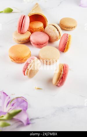 Assorted colorful macarons scattered with an empty ice cream cone and flowers on a white marble background. Stock Photo