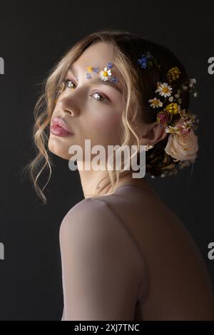 Young woman with long, blonde, curly hair, adorned with flowers in her hair and on her face, poses for a studio portrait. Stock Photo