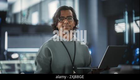 Young Indian Software Engineer Using Laptop Computer in Data Center, Looking at Camera. Successful Information Technology Manager Standing in a Room in a Tech Office Stock Photo