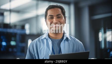 Handsome Smiling IT Specialist Using Laptop Computer in Data Center, Looking at Camera. Successful South Asian Businessman and e-Business Entrepreneur in Software Development Agency Stock Photo