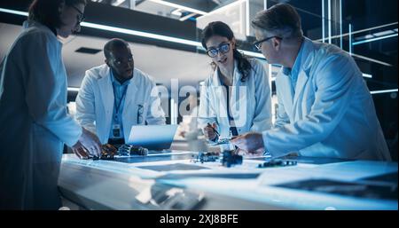 Industrial Robotics Engineers Working on a Humanoid Robot, Consulting with Specialists, Blueprints and Analyzing Electronic Components. Young Female Scientist Having a Meeting with Her Team Stock Photo