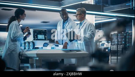 Diverse Team of Industrial Robotics Specialists Gathered Around a Table With a Mobile Robot. Engineers In Lab Coats Discussing Automated Robotic Delivery Assistant with Artificial Intelligence Stock Photo