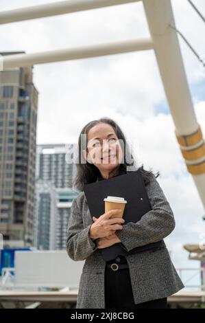 A confident, cheerful adult Asian businesswoman in a formal business suit stands in the urban city with a takeaway coffee cup and a document folder in Stock Photo