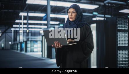 Muslim Female Data Center IT Engineer Standing in a Room with Server Racks. Cloud Computing Architect Uses Laptop Computer for Servicing the System in Cyber Security and Data Protection Facility Stock Photo