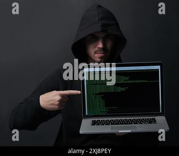 Crime, hoodie and hacker in studio, laptop screen and fraud of cybersecurity, online and pointing. Black background, scam and coding of criminal Stock Photo
