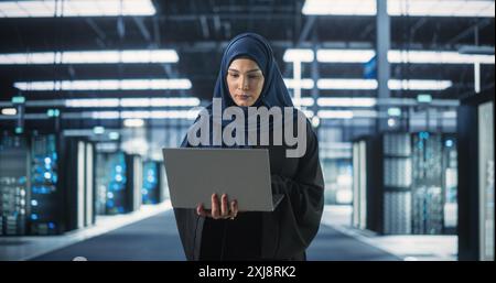 Muslim Female Data Center IT Engineer Standing in a Room with Server Racks. Cloud Computing Architect Uses Laptop Computer for Servicing the System in Cyber Security and Data Protection Facility Stock Photo