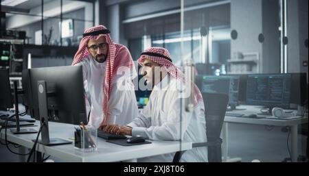 Two Young Saudi Software Engineers Use Desktop Computer to Discuss a Technological Project in a Modern Industrial Office. Arab Scientists Work in Research and Development Center Stock Photo