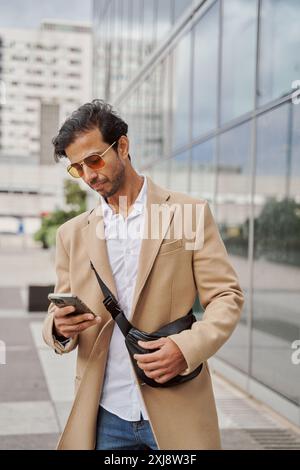 Happy handsome young middle eastern guy in casual and sunglasses going to office, walk by street, using phone, checking email, using business mobile a Stock Photo