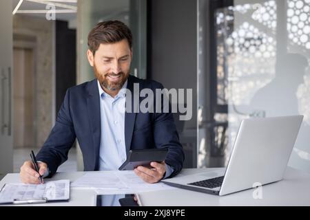 Smiling young male accountant, financial expert and analyst sitting in modern office at desk, working with documents and calculator. Stock Photo