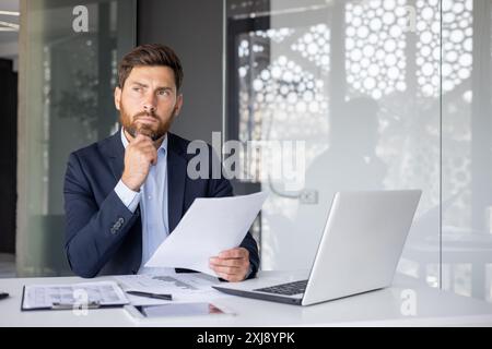 A serious and focused young man businessman works in the office, sits at a desk with a laptop, holds documents in his hands and looks thoughtfully to the side. Stock Photo