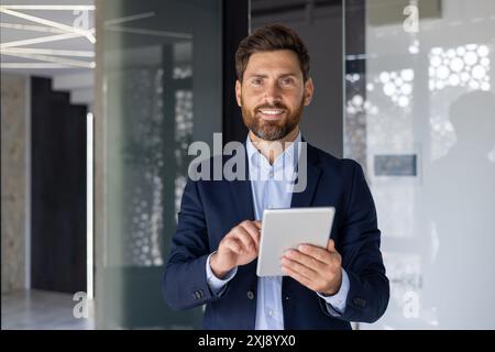 A professional businessman stands confidently in a sleek, modern office, holding a digital tablet, suggesting innovation and business efficiency. Stock Photo