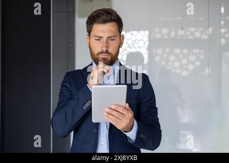 A focused mid-aged businessman in a suit critically analyzes data on his tablet within a modern office setting. He portrays concentration and professionalism. Stock Photo