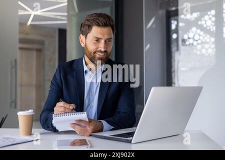 Professional middle-aged businessman in a modern office. Engaged in work, he uses a laptop and writes notes with a smiling demeanor. Stock Photo