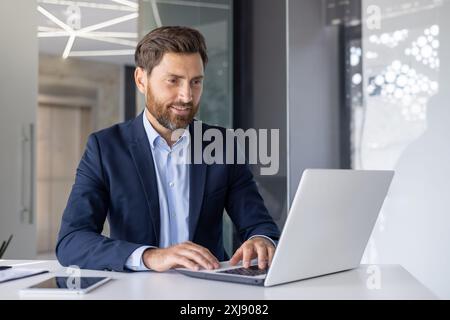Confident middle-aged businessman focused on working with a laptop in a bright, contemporary office setting. He appears engaged and serious about his task. Stock Photo