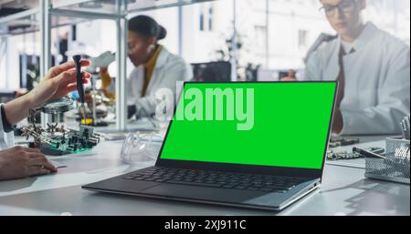 Laptop Computer with Green Screen Mock Up Display Standing on a Table in a Science Class Laboratory. Diverse Students Practicing Soldering Electronic Components for Their Tech Projects Stock Photo
